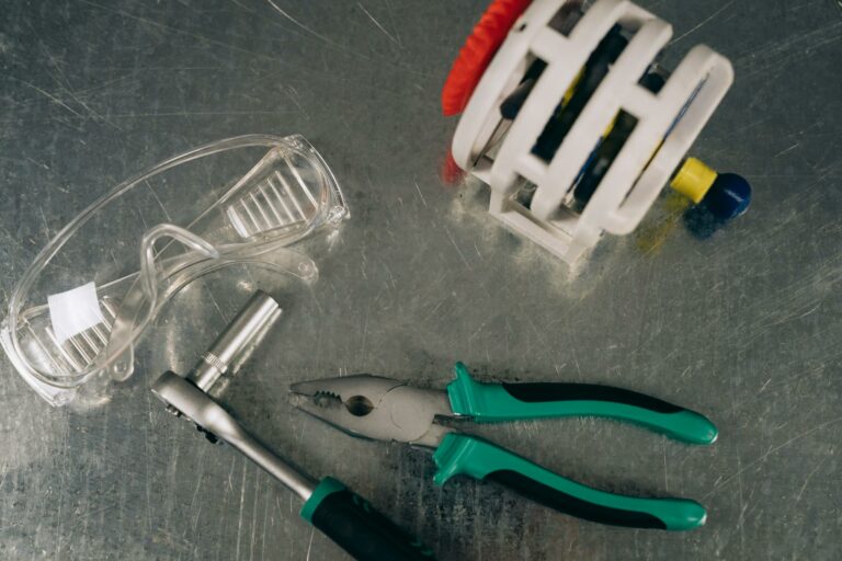 A flat lay of tools including pliers, safety glasses, and a ratchet on a shiny metal surface.