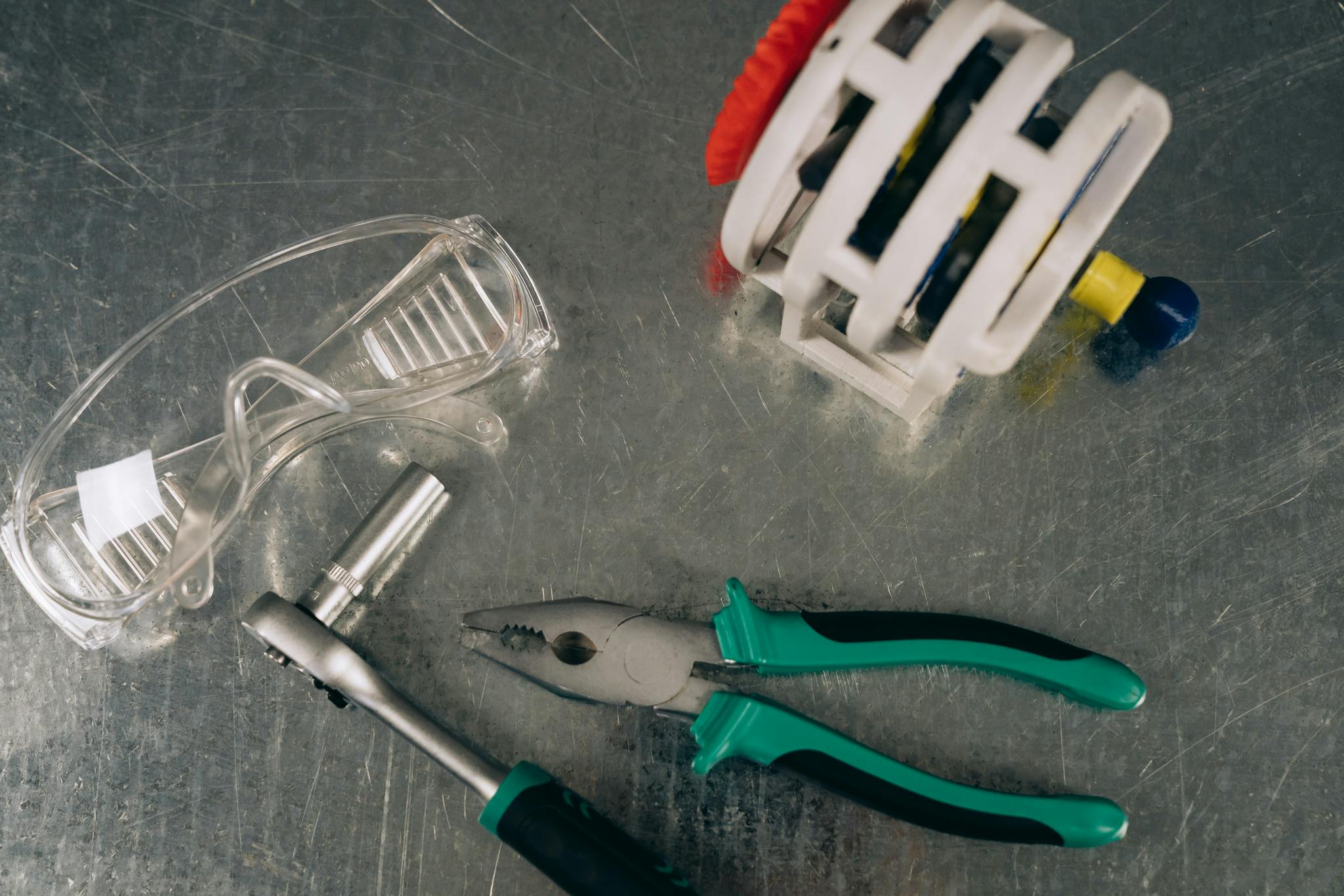 A flat lay of tools including pliers, safety glasses, and a ratchet on a shiny metal surface.