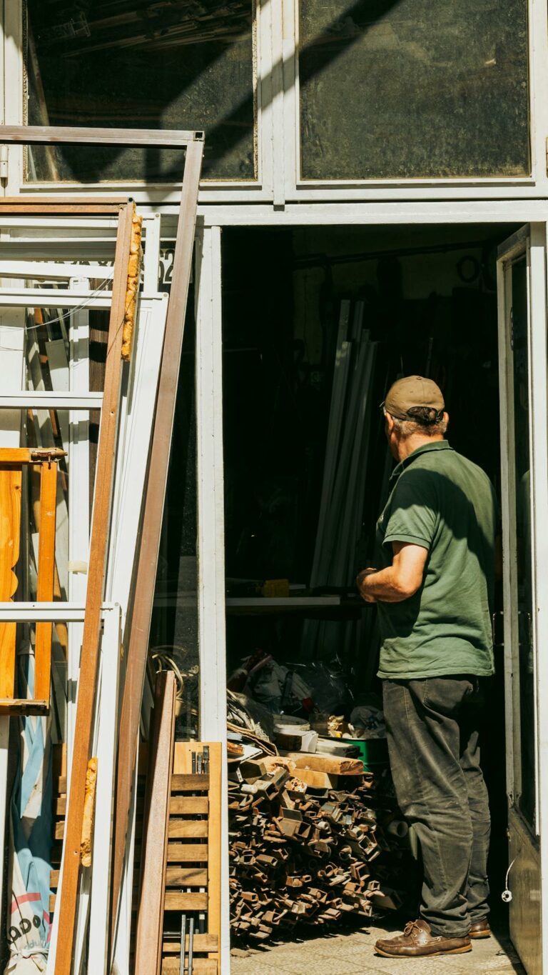 A man stands in a workshop shop filled with scrap items, captured in bright sunlight.